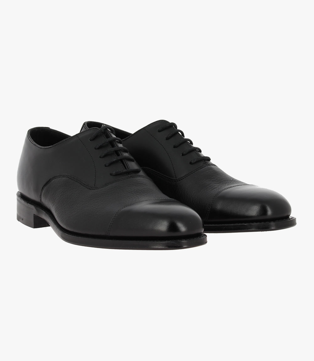 This dynamic image features both shoes positioned at an angle, allowing for a view of their symmetrical design and polished surfaces. The shoes are elegantly paired, highlighting their classic Oxford style. The subtle variations in light reflect off the leather, showcasing its rich texture and premium quality.