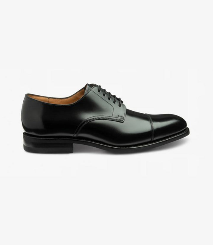 This side view captures the sleek silhouette of the Loake 305 Black Oxford Toe Cap Shoe, showcasing its elegant lines and refined toe-cap design. The polished black calf leather gleams under the light, highlighting the craftsmanship and attention to detail. The Goodyear welted rubber sole is visible, emphasizing durability and comfort. The shoe's overall profile exudes a timeless sophistication, making it a perfect choice for formal occasions.