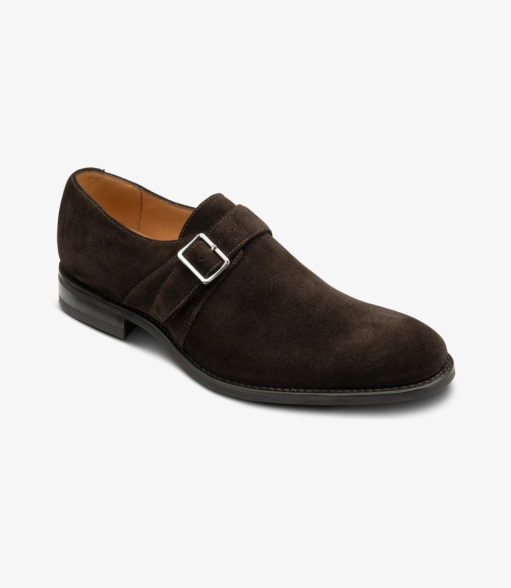 The shoe is displayed at an angle, showcasing the elegant curve of the toe cap, the secure monk strap fastening, and the suede's rich, velvety finish. The heel and sole structure are subtly visible, emphasizing both style and functionality.