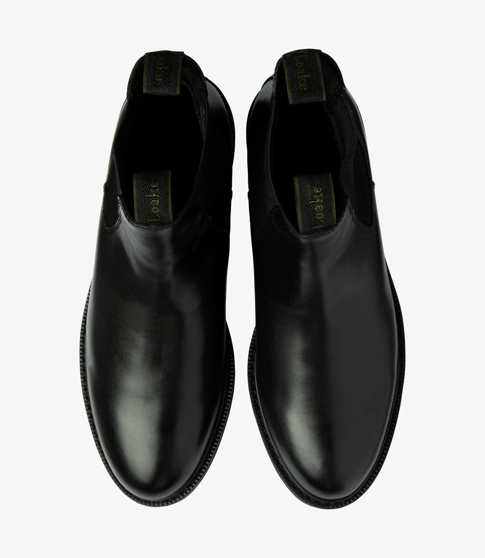 From above, the two Loake Ducksworth Black Chelsea Boots are displayed side by side, revealing their symmetrical design and sleek outline. The glossy surface of the waxy leather catches the light, while the round toe shape adds a classic touch. The interior of the boots is partially visible, showcasing the textile waterproof lining, emphasizing their practicality. The cemented rubber soles are also highlighted, indicating durability and traction, making these boots both stylish and functional.