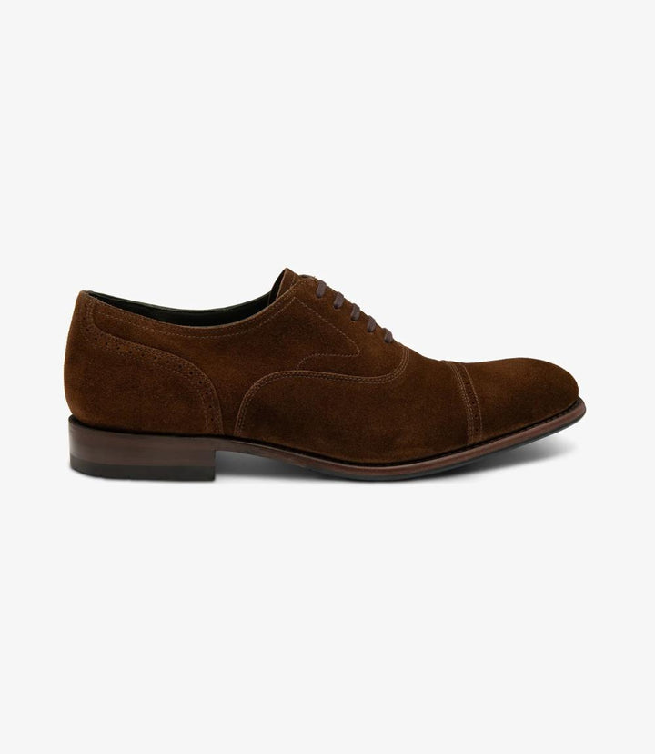 The photo showcases the Loake Hughes Brown Suede Oxford from the side, highlighting its elegant silhouette and the soft, textured suede material. The classic punched toe-cap design is prominently featured, along with the Goodyear welted sole that seamlessly combines leather and rubber for durability and style. The overall color is a rich brown, exuding sophistication.