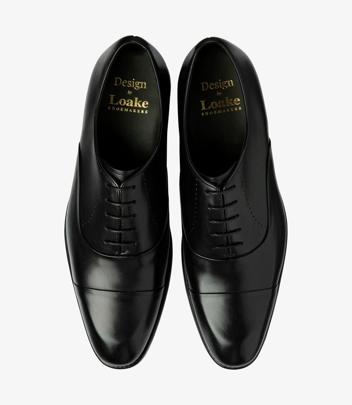 This overhead view features both shoes side by side, displaying their matching sleek designs. The polished black calf leather shines, while the toe-cap and stitching details are highlighted. The rubber soles provide a sturdy foundation, underscoring the shoes' practicality without compromising style. The overall look reflects sophistication and attention to detail.