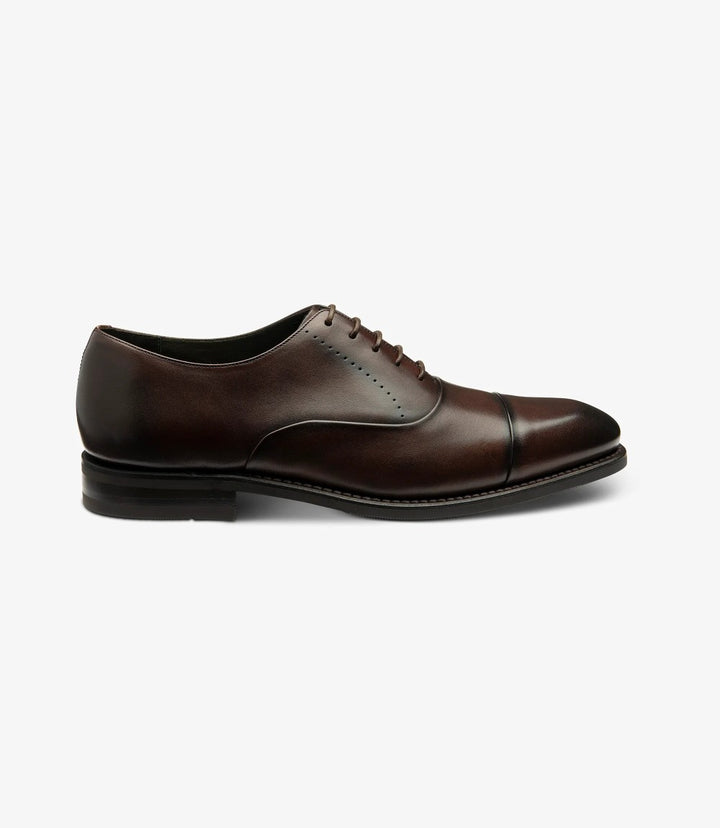 The image showcases the sleek profile of the Loake Phoenix Dark Brown Oxford Toe Cap. The polished hand-painted dark brown calf leather glistens under the light, accentuating the elegant toe-cap design. The Goodyear welted shadow rubber sole is visible, emphasizing the shoe’s robust construction and craftsmanship.