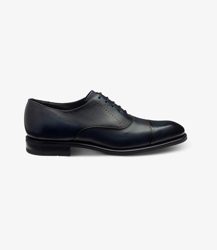 The photo showcases the Loake Phoenix Navy Oxford Toe Cap shoe from the side, highlighting its sleek silhouette and elegant design. The rich navy leather is complemented by a polished finish, while the structured toe cap adds a refined touch. The Goodyear welt construction is subtly visible, emphasizing the shoe's durability.