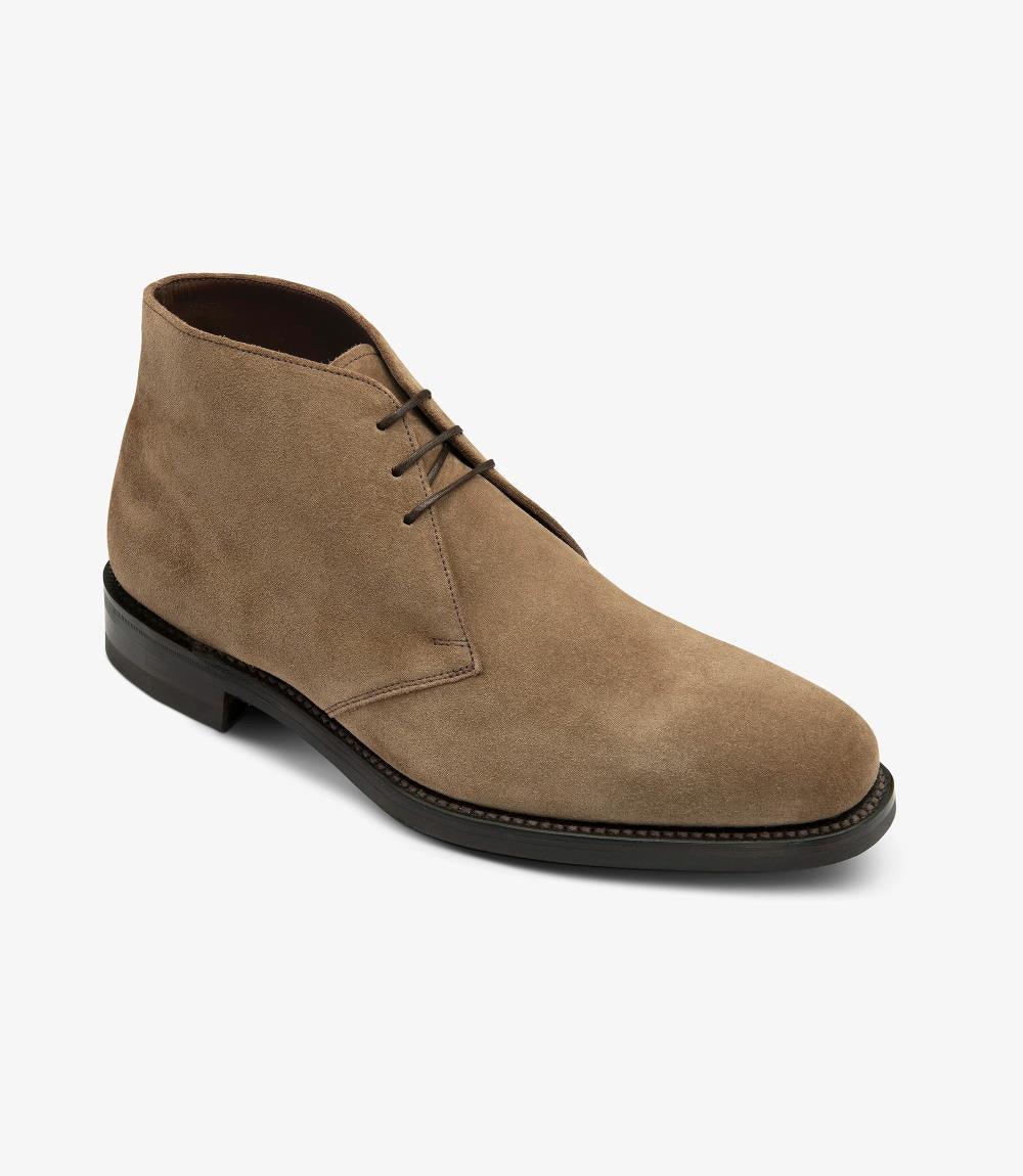 This angled shot of the boot captures its sophisticated chukka design. The rich flint suede is accentuated by the natural light, revealing subtle texture variations. The lace-up closure adds a classic touch, while the fully leather-lined interior emphasizes quality craftsmanship. The rubber sole is slightly visible, reinforcing the boot's rugged yet refined aesthetic.