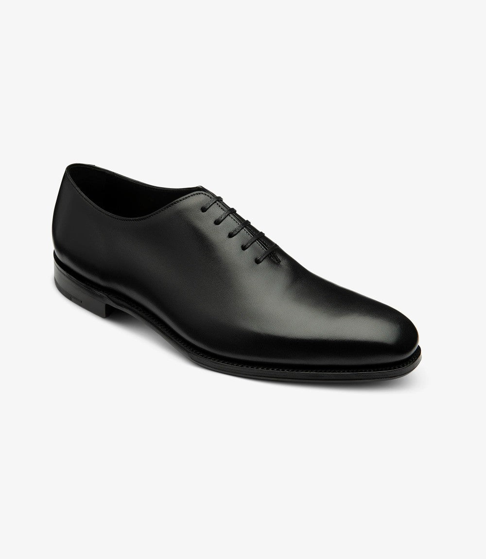 This angled view captures the Loake Wholebury Black Oxford Shoes in a dynamic perspective, highlighting the contours of the shoe. The polished black leather glistens under the light, revealing a subtle sheen that speaks to its premium quality. The toe cap features a delicate yet distinctive design, enhancing the overall elegance. The slightly elevated heel is visible, adding to the shoe's profile and ensuring comfort without compromising style.