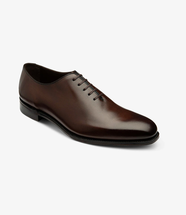 From this angled perspective, the Loake Wholebury Dark Brown Oxford Shoes reveal their sleek profile and refined detailing. The polished dark brown leather glistens under the light, accentuating the shoe's luxurious appearance. The rounded toe adds a timeless touch, while the slightly elevated heel contributes to an elegant stance. The laces are securely tied, and the shoe’s contours are smoothly defined, showcasing the perfect blend of style and comfort. 