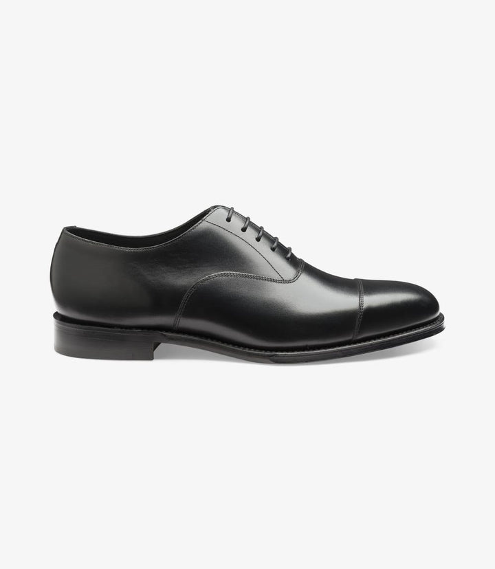 The photo showcases the sleek side profile of the Loake Aldwych Black Oxford Toe-Cap shoes. The polished black calf leather gleams under the light, highlighting the sophisticated toe cap design. The smooth silhouette is complemented by the classic lace-up closure, while the Goodyear welted leather sole is visible, emphasizing the shoe's quality construction.