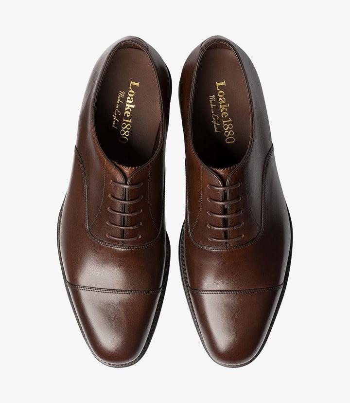 The overhead view reveals the complete outline of the Aldwych Oxford shoes, showcasing the symmetrical toe-cap design. The polished surface of the mahogany leather gleams under the light, while the smooth lines converge towards the sleek laces. This angle highlights the shoe's elegant profile and craftsmanship, giving a clear view of the leather texture and quality construction.