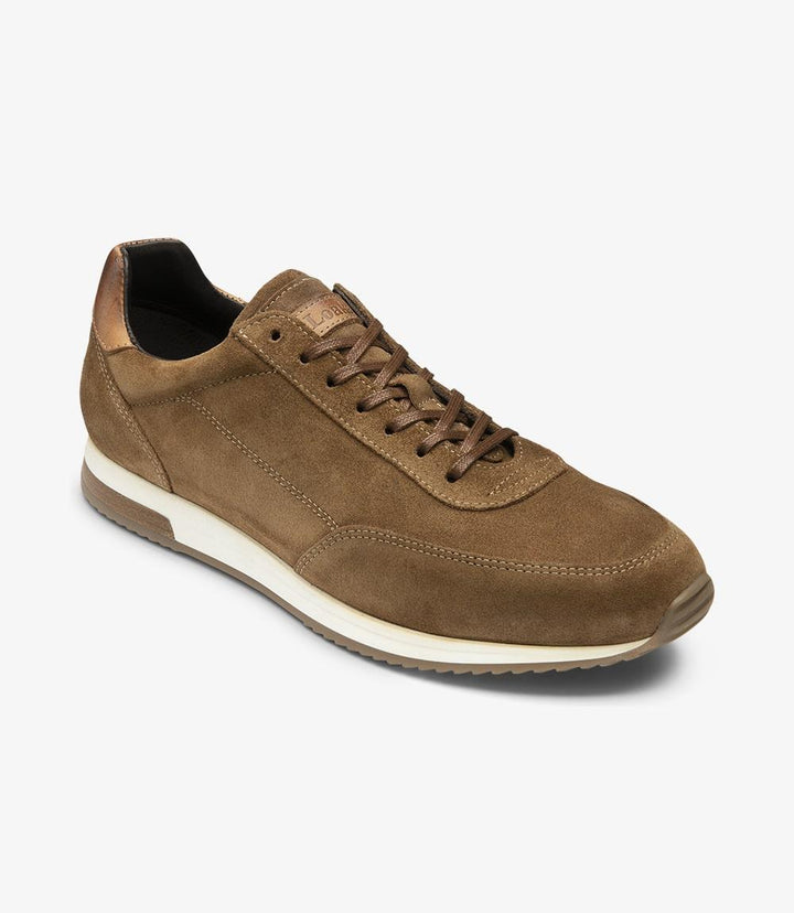 In this angled side view, the Loake Bannister Tan Suede Trainer reveals its distinctive toe shape and the curvature of the shoe. The suede upper appears soft and inviting, with the light reflecting off its texture, highlighting the craftsmanship. The contrasting rubber sole is more prominent, showcasing its grip and durability. This perspective emphasizes the shoe's modern design while maintaining a classic appeal, perfect for casual or semi-formal occasions.