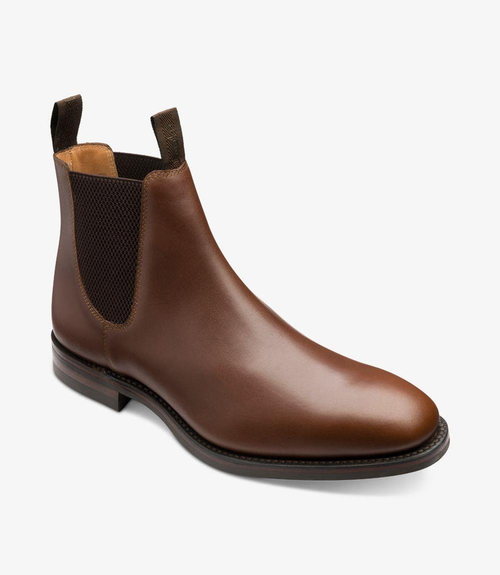 Captured from a dynamic angle, this image emphasizes the boot's streamlined shape and the beautiful curvature of the upper. The light catches the waxed leather, revealing subtle variations in color and texture that enhance its sophistication. The padded collar adds a hint of comfort, making it perfect for all-day wear. The Dainite sole's tread pattern is partially visible, illustrating the boot's traction capabilities.