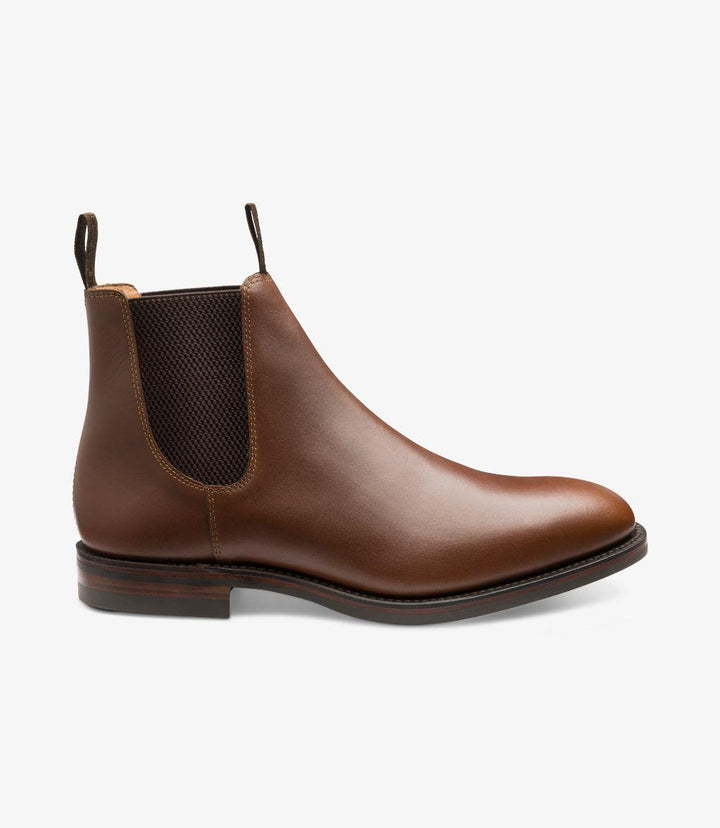 The side view of the Loake Chatsworth Chelsea Boot showcases its sleek silhouette and elegant lines. The rich brown waxed leather glistens under the light, highlighting its premium quality. The elastic side panel, designed for easy on and off, seamlessly integrates with the boot's design. The Dainite rubber sole is visible at the bottom, providing a sturdy foundation while adding a contemporary touch.