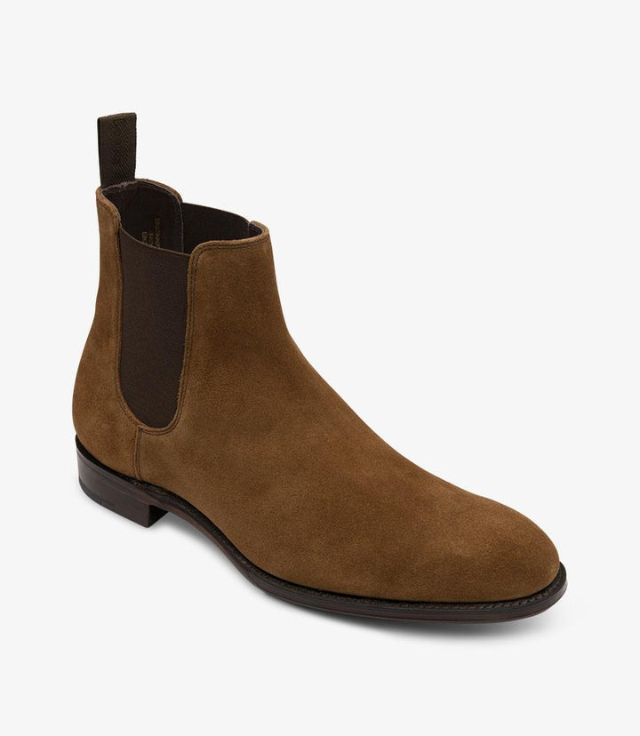 This angled view captures the right side of the Loake Coppergate Tobacco Suede Chelsea Boot. The perspective emphasizes the curvature of the boot and its streamlined shape. The subtle sheen of the eco suede is evident, reflecting light softly. The photo also highlights the contrast between the deep tobacco color and the leather sole, showcasing the boot's premium materials and craftsmanship.