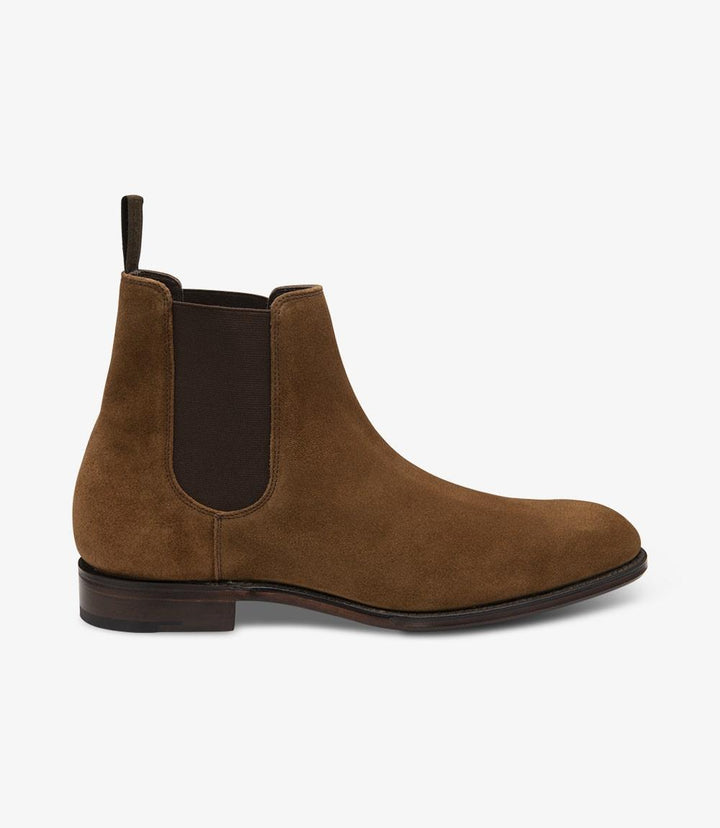 The photo showcases the left side of the Loake Coppergate Tobacco Suede Chelsea Boot. The rich tobacco suede is prominently displayed, highlighting its soft texture and elegant finish. The boot features a sleek silhouette with a slightly tapered toe, complemented by the classic elastic side panels that enhance its Chelsea boot design. The Goodyear welted leather sole is visible at the base, indicating its robust construction.