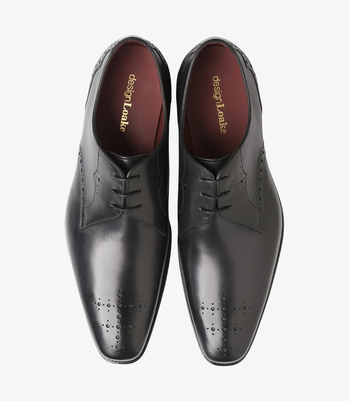 The photograph captures both shoes from above, arranged side by side. The polished black leather surfaces shine brightly, revealing the meticulous hand-painted finish. The symmetry and balance of the design are evident, showcasing the classic Derby shape. The punched detailing around the toe area adds a touch of sophistication, while the sleek lines create an appealing aesthetic.