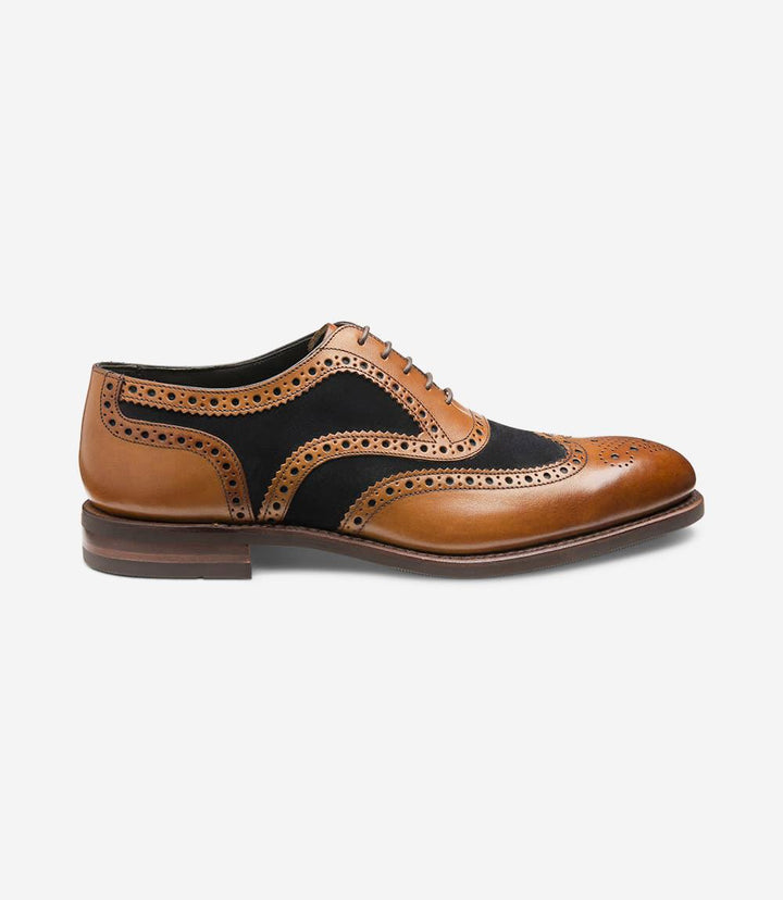 The Loake Kerridge shoe features a sophisticated tan and navy color combination in premium calf leather. The side showcases intricate brogue detailing along the seams, with small punched holes forming classic wingtip and spider patterns. The slightly rounded toe cap is elegantly defined, while the rubber 'Shadow' sole is visible at the base, offering a subtle contrast. The shoe has a low stacked heel and a fine welt that emphasizes its polished look.