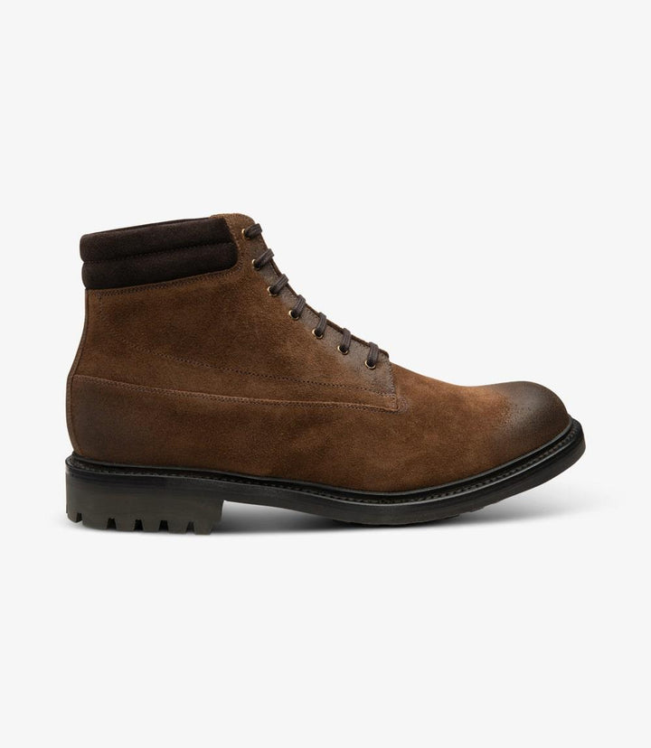 This image showcases the side profile of the Loake Kirkby Brown Suede Boot. The soft suede leather is richly textured, presenting a warm brown hue that exudes sophistication. The boot features a classic lace-up design with strong, sturdy eyelets and flat laces, providing a secure fit. The padded collar around the ankle adds an element of comfort and support. The Goodyear welted commando rubber sole is clearly visible, highlighting its rugged durability and traction.