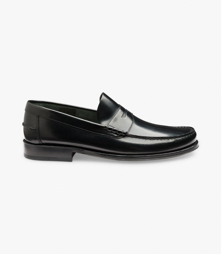 The side view of the Loake Princeton Black Loafer showcases its sleek and polished silhouette. The black leather gleams in the light, highlighting the subtle grain texture. The elegant beef-roll detailing runs along the top of the shoe, adding a touch of sophistication. The rounded toe shape enhances its classic design, while the flexible leather sole curves gently, reflecting the loafer's comfortable and stylish construction. 