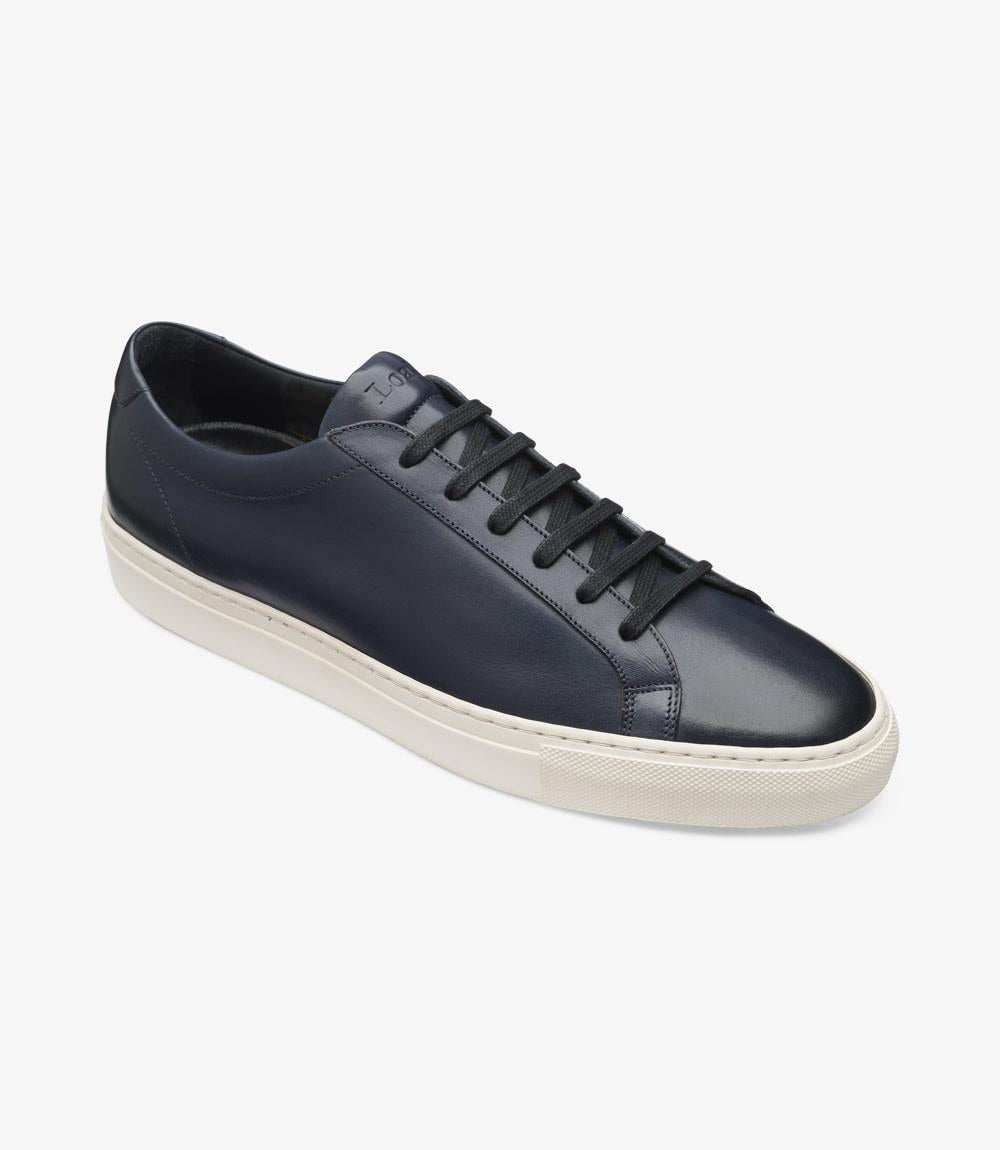 In this angled view, the Loake Sprint Navy Sneaker reveals its dynamic profile, highlighting the bold toe and generous fit offered by the sneaker last shape. The hand-painted navy leather glistens under the light, reflecting its high-quality craftsmanship. The rubber cup sole is visible, emphasizing its thickness and traction features. This angle captures the essence of versatility and comfort, making it suitable for various occasions.