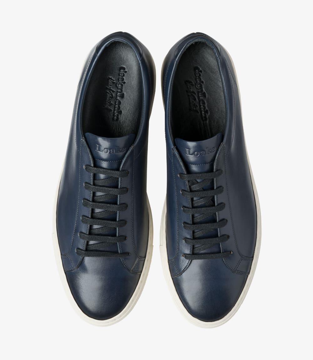 From above, the Loake Sprint Navy Sneaker displays its clean lines and refined shape. The classic rounded toe design is prominent, while the lacing system is neatly positioned at the center, providing a snug fit. The navy leather appears rich and luxurious, while the smooth surface showcases the attention to detail in its craftsmanship. The overall symmetry of the sneaker emphasizes its contemporary style.