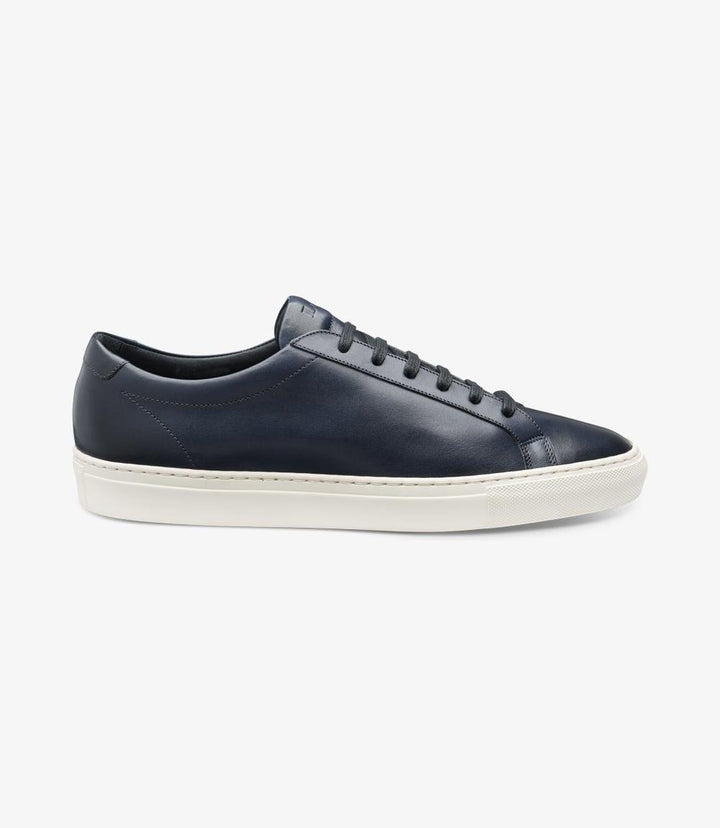 The side view of the Loake Sprint Navy Sneaker showcases its sleek silhouette and premium hand-painted navy calf leather. The smooth leather upper is complemented by a subtly contoured design that flows seamlessly into the sturdy side-stitch rubber cup sole, which adds a sporty touch while providing durability. The rounded toe shape enhances the shoe's modern aesthetic, making it a stylish choice for casual or smart-casual wear.