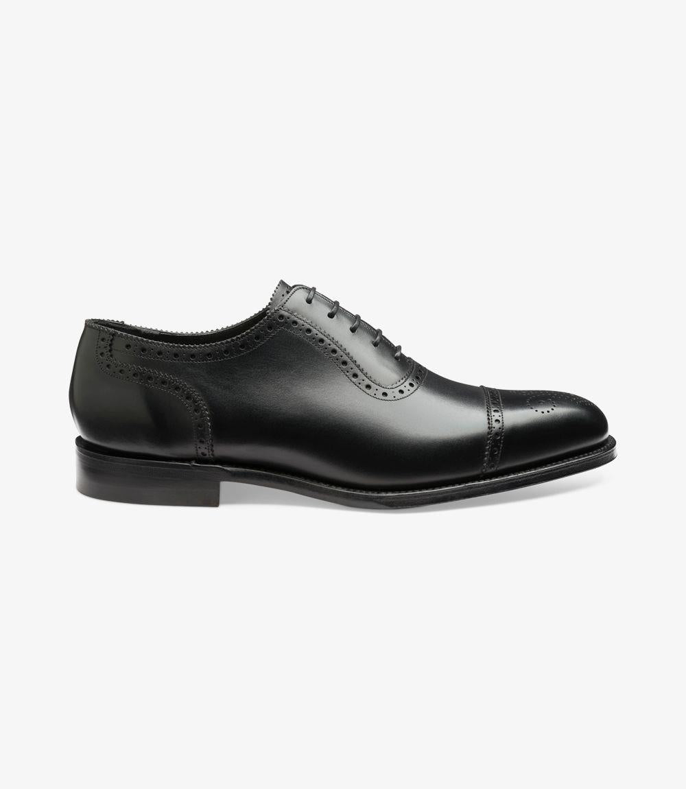 The side profile of the Loake Strand Black Semi-Brogue shoe showcases its elegant lines and sophisticated silhouette. The burnished calf leather gleams subtly in the light, while the brogue detailing around the toe cap adds a refined touch. The Goodyear welted sole is visible, emphasizing the shoe’s durable construction.