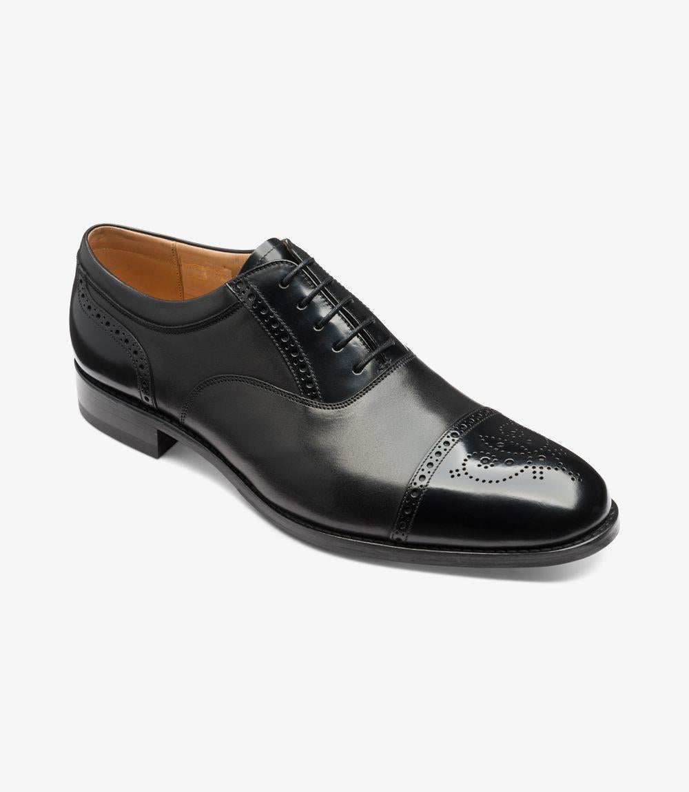 This angled perspective captures the Loake Woodstock Black Oxford Semi Brogue in a dynamic light, showcasing its sophisticated profile. The polished black leather exudes elegance, while the rounded toe provides a classic touch. The semi brogue detailing adds character to the shoe, with its tasteful perforations and decorative elements clearly visible from this angle. The slightly raised heel contributes to the shoe's refined look, making it perfect for formal settings. 